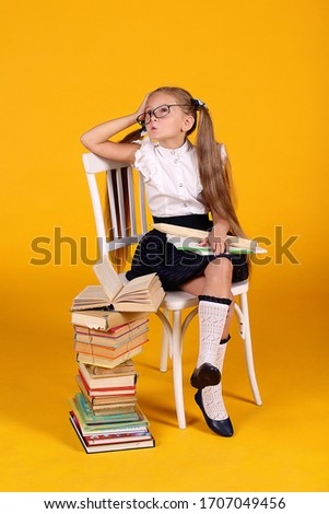 Girl student first-grader in a school uniform and glasses sits on a white chair, next to a stack of books. In the hands of books and large pencils. Yellow background.