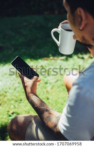 Mockup shot of man's hand with tattoos holding  cell phone and coffee cup with blank screen in backyard.