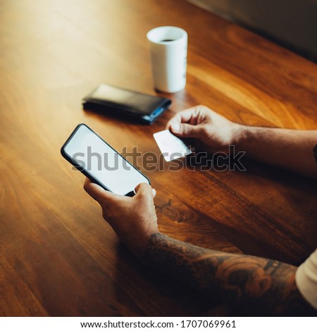 Mockup shot of man's hand with tattoos holding  cell phone and credit card with blank screen on desk at home office. For shopping.