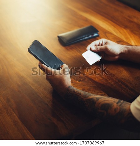 Mockup shot of man's hand with tattoos holding  cell phone and credit card with blank screen on desk at home office. For shopping.