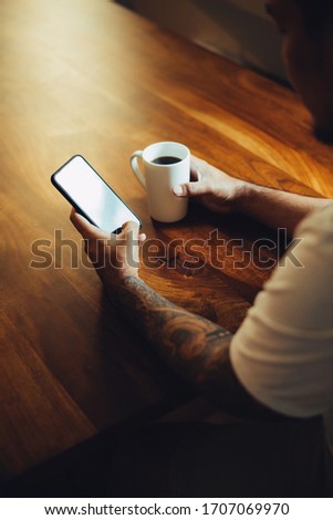 Mockup shot of man's hand with tattoos holding  cell phone and coffee cup with blank screen on desk at home office.
