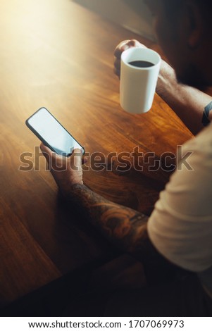 Mockup shot of man's hand with tattoos holding  cell phone and coffee cup with blank screen on desk at home office.