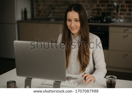Remote work. A caucasian brunette woman with headphones working remotely on her laptop. A happy girl in a white shirt doing business at her home workplace.