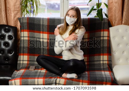 Portrait of a cute caucasian girl in a medical mask in a white sweater sitting in front of the camera. Model posing on the sofa at home.