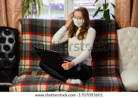 Portrait of a cute caucasian girl in a medical mask in a white sweater sitting in front of the camera. The model works on a laptop on the sofa in a home setting.