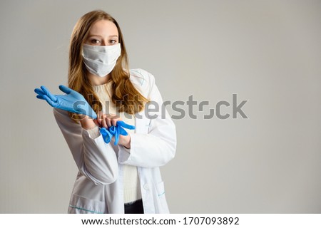 Studio portrait of a pretty blonde girl doctor in a protective mask and white coat. A woman is standing right on a white background in protective gloves.