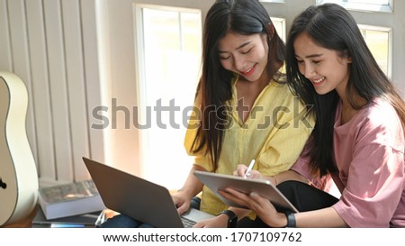 Photo of two beautiful women relaxing with computer laptop and tablet while sitting together at the floor over comfortable music studio as background.