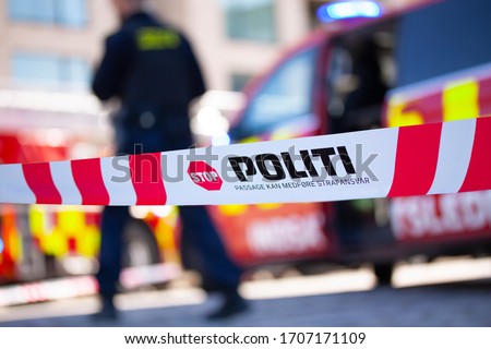 Shallow depth of field image of police tape in Denmark with blurred danish police man and emergency vehicle in the background. Text on tape saying Police line do not cross.