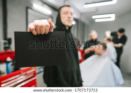 Barber in black hoodie holds black card on barber shop background and hairdressers clipping clients. Man holds a blank card in his hand on a background of a male hairdresser.