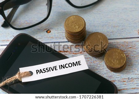 Top view of stack of gold coins,glasses,mobile phone and paper tag written with Passive income on wooden background.