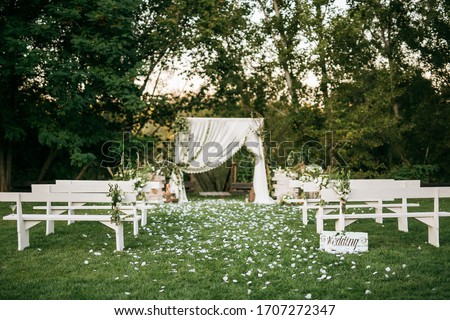 Wedding ceremony aisle with an arch made of chiffon white textile and flowers and long white wooden benches on green grass. Backyard wedding venue. Royalty-Free Stock Photo #1707272347