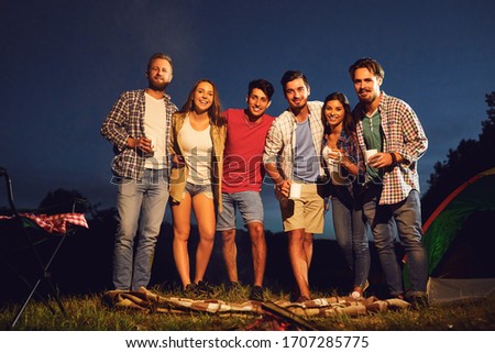 A group of people standing by the bonfire next to the tent at night in the summer in autumn.