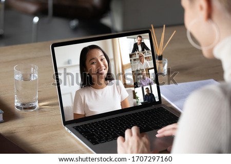 Asian woman and diverse colleagues taking part in group videocall laptop screen view over female shoulder sitting at desk working from home. Distant chat, virtual communication, video call app concept Royalty-Free Stock Photo #1707297160