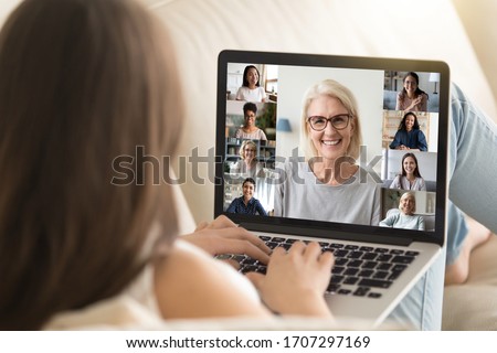Laptop screen view mature 50s lady lead group videocall distant talk with different ethnicity age women. View over girl shoulder sit on sofa involved at remote chat, empowerment movement club concept