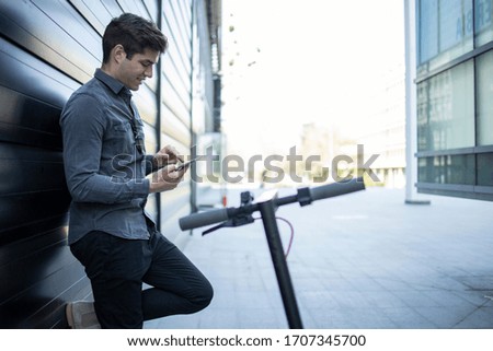 Business person by his electric scooter checking email on his tablet computer on the street. Business buildings exterior in background.