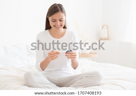 Young lady feeling happy about getting pregnant that she learnt from pregnancy test she is holding while sitting on bed