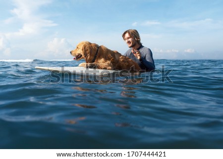 Surfing. Handsome Surfer With Dog On Surfboard Swimming In Ocean. Waved Water Surface, Blue Sky With Soft Clouds On Background.