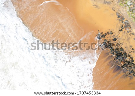 aerial drone bird view shot of the sea shore with yellow sand, black rocks, large white waves and foam crashing on the beach forming beautiful textures, patterns, shapes. Sri Lanka