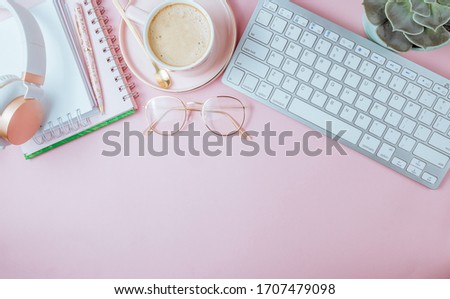 Flat lay of office feminine desk workspace with cup of coffee, succulent, laptop, empty notebook, headphones, keyboard, and golden eyeglasses on pink background. Top view. Copy space