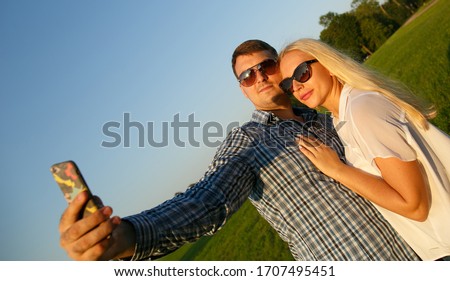 Cheerful and happy newlyweds in their honeymoon. Emotional portrait of a positive and tanned blonde girl hugging her beloved husband during a joint photo on a smartphone on a walk in the park. Selfies