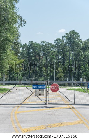 Closed entrance to park and nature preserve in suburban Houston, Texas, America. Park closed with Stop side on metal fence barricade. Row of matured pine trees in background.