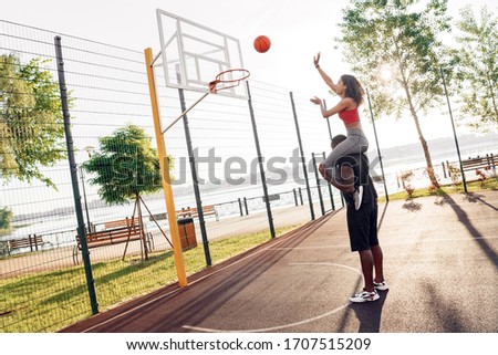 Young african descent couple girlfriend sitting on shoulders of boyfriend standing on basketball court outdoors throwing ball to basket excited