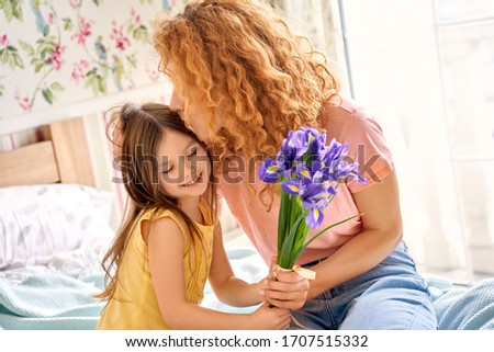 Birthday concept. Young adult mother cuddle and kissing her happy kid girl, holding flowers in hand, sitting in bedroom