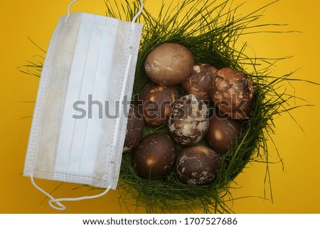 multi-colored Easter eggs in a nest of green grass, with a medical mask against viruses, on a yellow background