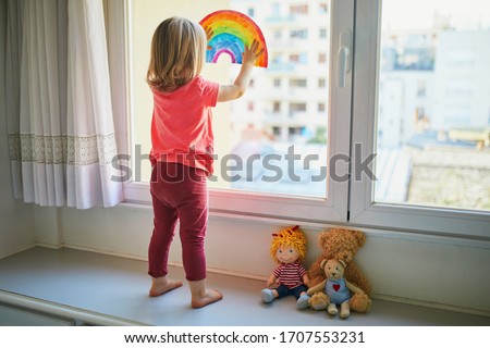 Adorable toddler girl attaching rainbow drawing to window glass as sign of hope. Creative games for kids staying at home during lockdown. Self isolation and coronavirus quarantine concept