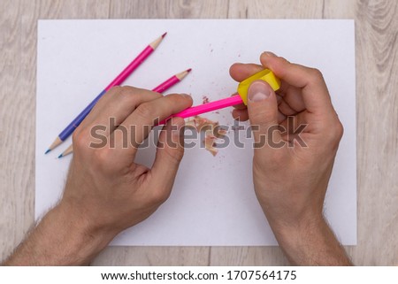 the man's hands are sharpened with a sharpener a pink pencil on a white sheet next to which are shavings from a pencil and colored pencils scattered