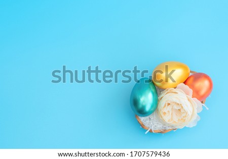 Decorated Easter eggs lie in the basket like a nest with white flower on blue background. Happy Easter holiday concept. Greeting or invitation card. Flat lay style with copy space.