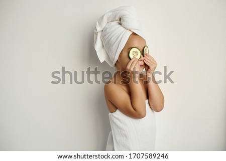 A young caucasian child with wrapped hair in white bath towels applying pieces of cucumber to her eyes on a white background