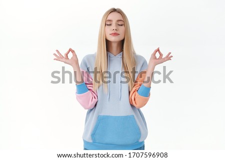 Relaxed, peaceful blonde girl in hoodie, keep calm and patient, close eyes and breathing during meditation, reach zen or nirvana, hold hands sideways, standing in lotus pose white background