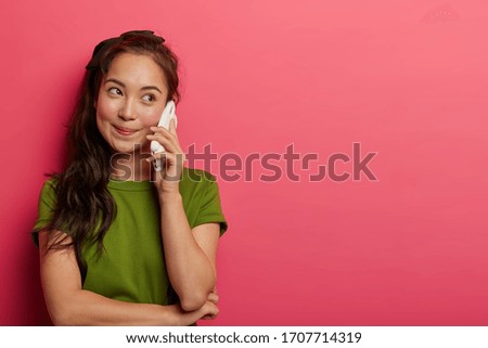 Pleasant looking natural brunette girl holds smartphone near ear, enjoys nice phone talk, looks aside, wears casual t shirt, discusses something interesting with friend, isolated on pink background