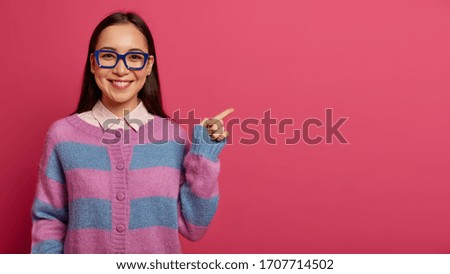 Beautiful brunette Asian woman indicates aside, shows promo, gives advice, makes choice, smiles pleasantly, wears transparent glasses, stands against rosy studio wall, tells about product details Royalty-Free Stock Photo #1707714502