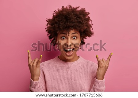 Fun and amusement. Joyful dark skinned young woman shows heavy metal gesture, foolishes around at concert, exclaims gladfully as hears favorite rock song, enjoys good loud sound, stands indoor