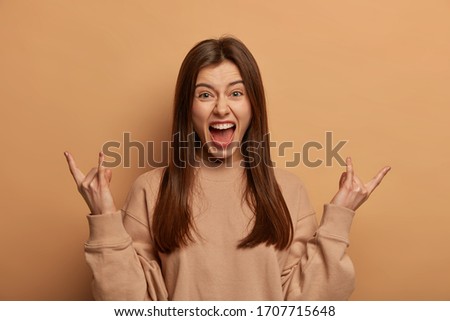 Emotive brunette woman makes heavy metal sign, rock n roll gesture, gestures actively, attends concert of favorite band, expresses positive emotions, wears sweatshirt, models over beige wall