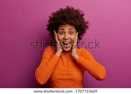 Happy energetic woman grabs face, hears wonderful news, realises she won huge money, stands impressed and speechless, dressed in orange jumper, feels lucky and thrilled, got incredible offer Royalty-Free Stock Photo #1707715669