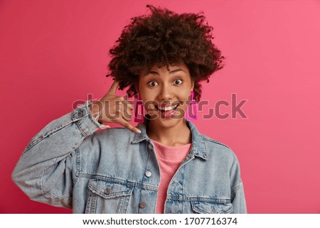 Young fashionable woman makes telephone gesture, asks to call her, expresses positive emotions, wears denim clothes, smiles happily, isolated on rosy background. Call me back sign. Body language
