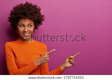 Good looking woman points right at copy space blank for your promotion, gives advice to click link, dressed in orange jumper, smiles pleasantly, isolated on bright purple wall recommends order product