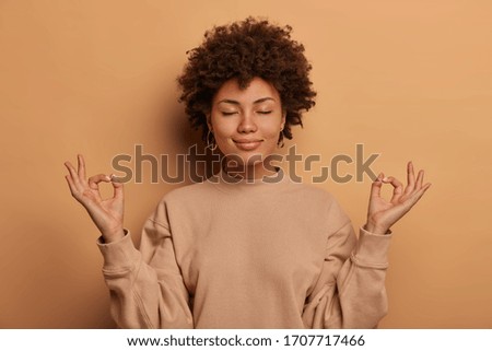Calm relieved dark skinned woman takes deep breath, keeps hands sideways in zen gessture, reaches nirvana and practices yoga, stands with closed eyes, stands stress free against brown background
