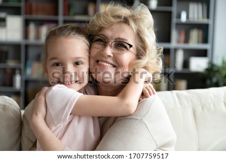 Close up of aged grandmother hugs little granddaughter people sitting on couch at home smile looking at camera. Multi-generational family portrait, candid feelings strong attachment and love concept