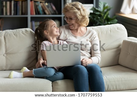 50s grandmother and small granddaughter sit on couch using laptop having fun watch cartoons online, different generations older younger people easy modern tech usage, educational program users concept