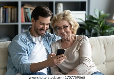 Millennial adult son demonstrates to older mother new application on smartphone sit on couch at home, helps her and teaches, shows old mom photos, share personal, having fun, modern tech usage concept Royalty-Free Stock Photo #1707759565