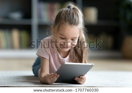 Little girl lying on floor in living room with tablet looking at device screen watching cartoons enjoy audiobook listening, preschool modern tech user parental control safety, gadget overuse concept