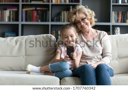 Multi generational family and modern tech, parental control, children protection in internet concept. Grandma sit on sofa with granddaughter holds cellphone use new app for kids, read e-books on-line