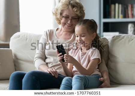 In living room sit on sofa resting little granddaughter with old grandma, kid holds cell phone. New generation addicted to wireless gadgets, website content parental control, bad habit overuse concept