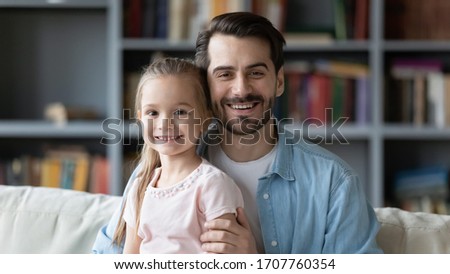Horizontal image head shot portrait young father and little daughter sitting on couch in cozy living room smiling looking at camera, capture moment for family album memories, happy fatherhood concept