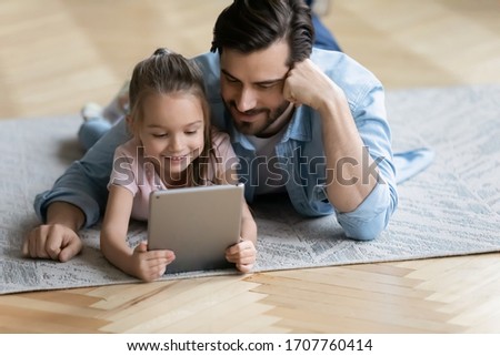 On carpet lying little daughter with father watching cartoons using tablet gadget. Warm floor modern home, devices overuse, e-books for kids on-line, parental control protect from bad content concept