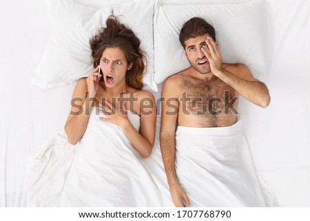 Shocked emotional woman hears bad news while talks via smartphone in bed, bored man lies near, tired of wife gossiping, needs attention, has uninterested expression, sleep comfortably in bedroom Royalty-Free Stock Photo #1707768790